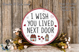 a round sign with the text "I WISH YOU LIVED NEXT DOOR" surrounded by a floral arrangement and a wooden background.