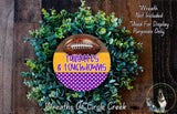 a wreath made of green foliage and a football, with the text "Tailgates & Touchdowns" written on it. The wreath is displayed on a wooden surface, and there is a small dog in the bottom right corner of the image.