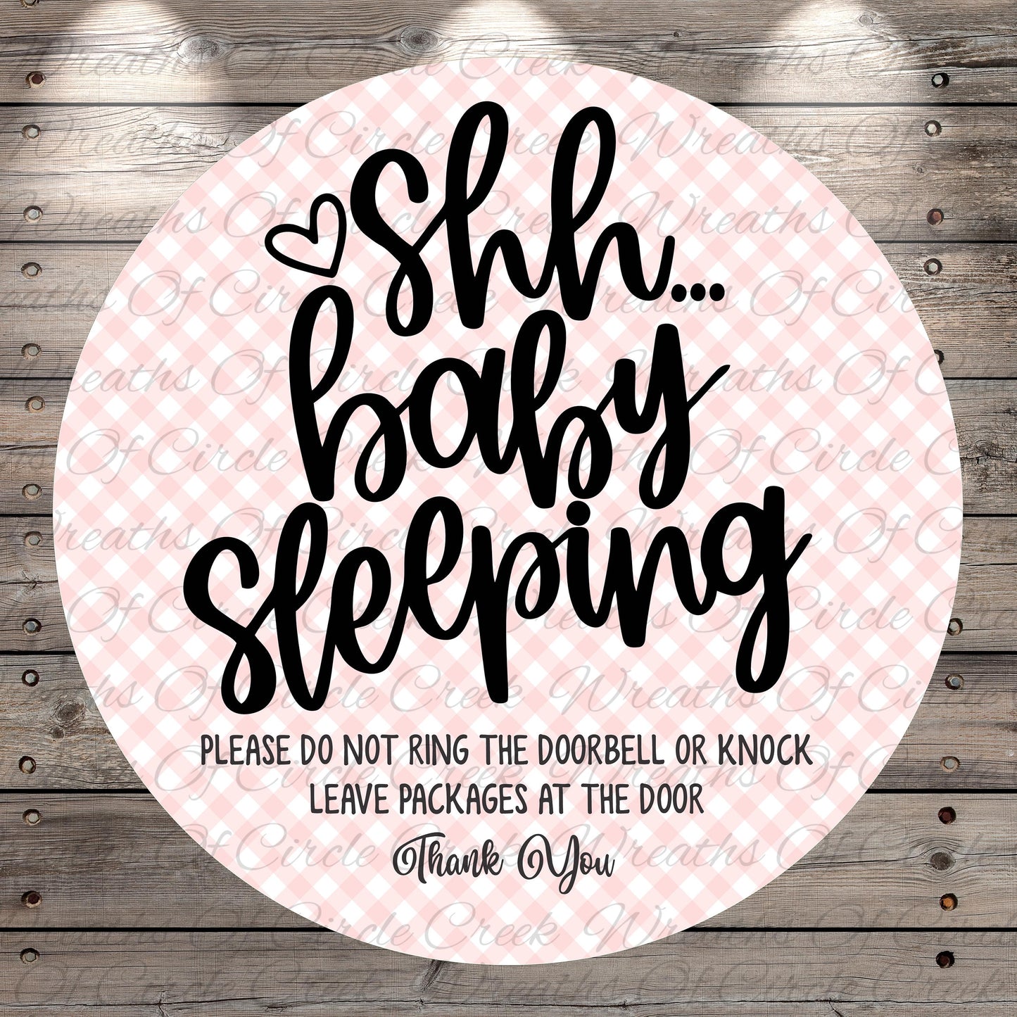 a circular sign with a pink and white checkered background and black text. The sign reads "Shh... baby sleeping" and includes a warning that says "PLEASE DO NOT RING THE DOORBELL OR KNOCK LEAVE PACKAGES AT THE DOOR Thank You".