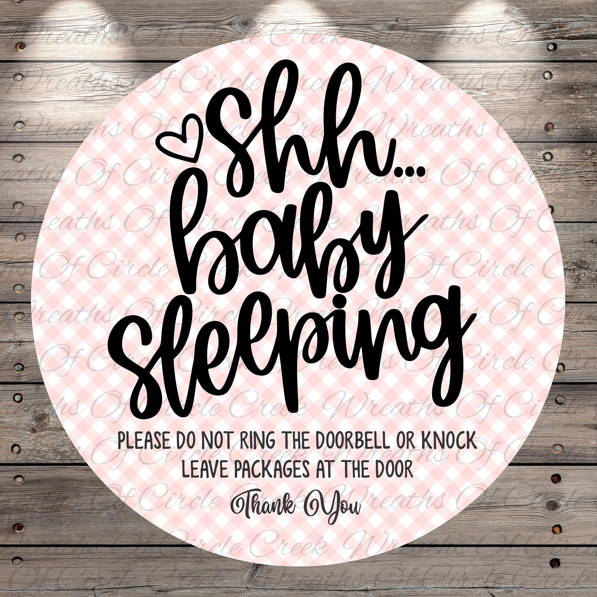 a circular sign with a pink and white checkered background and black text. The sign reads "Shh... baby sleeping" and includes a warning that says "PLEASE DO NOT RING THE DOORBELL OR KNOCK LEAVE PACKAGES AT THE DOOR Thank You".
