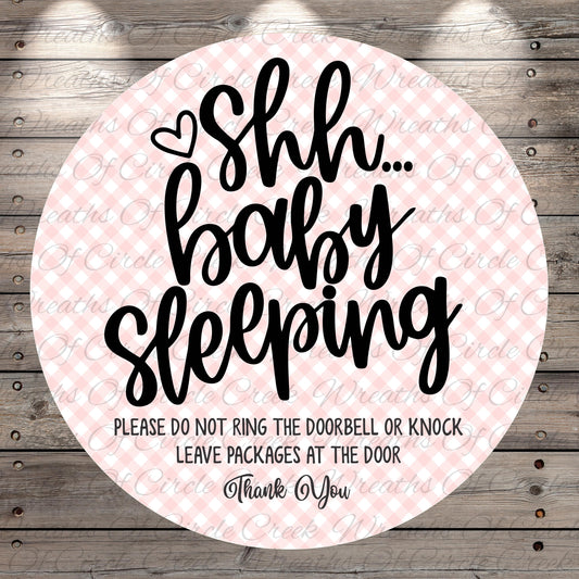 a circular sign with a pink and white checkered background and black text. The sign reads "Shh... baby sleeping" and includes a warning that says "PLEASE DO NOT RING THE DOORBELL OR KNOCK LEAVE PACKAGES AT THE DOOR Thank You".
