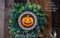 A Halloween-themed wreath made of green foliage and a carved pumpkin with a smiling face, placed on a wooden surface.