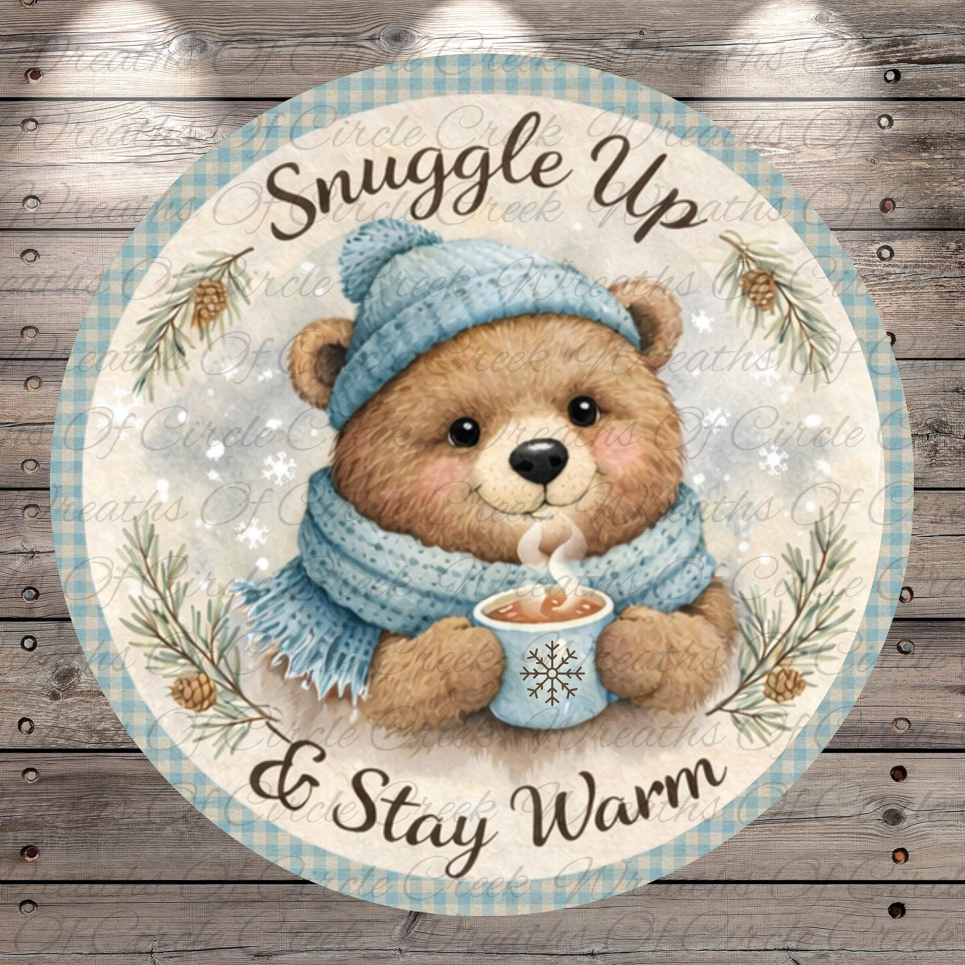 A round, decorative plate with a teddy bear wearing a blue hat and scarf, holding a cup of hot chocolate, and surrounded by pine branches and snowflakes. The text on the plate reads "Snuggle Up & Stay Warm".