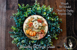 a wreath made of various green plants and flowers, including pumpkins and other fall-themed elements, displayed on a wooden surface.