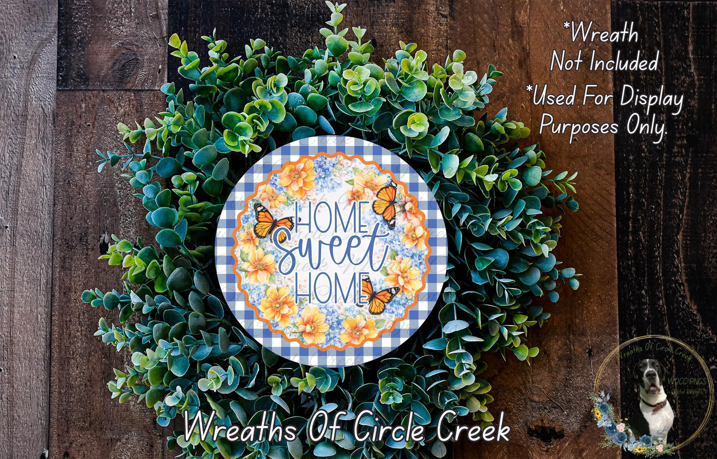 a wreath made of various green plants and flowers, with a decorative plate in the center that says "Home Sweet Home". The wreath is displayed on a wooden surface, and there is a small dog in the bottom right corner.