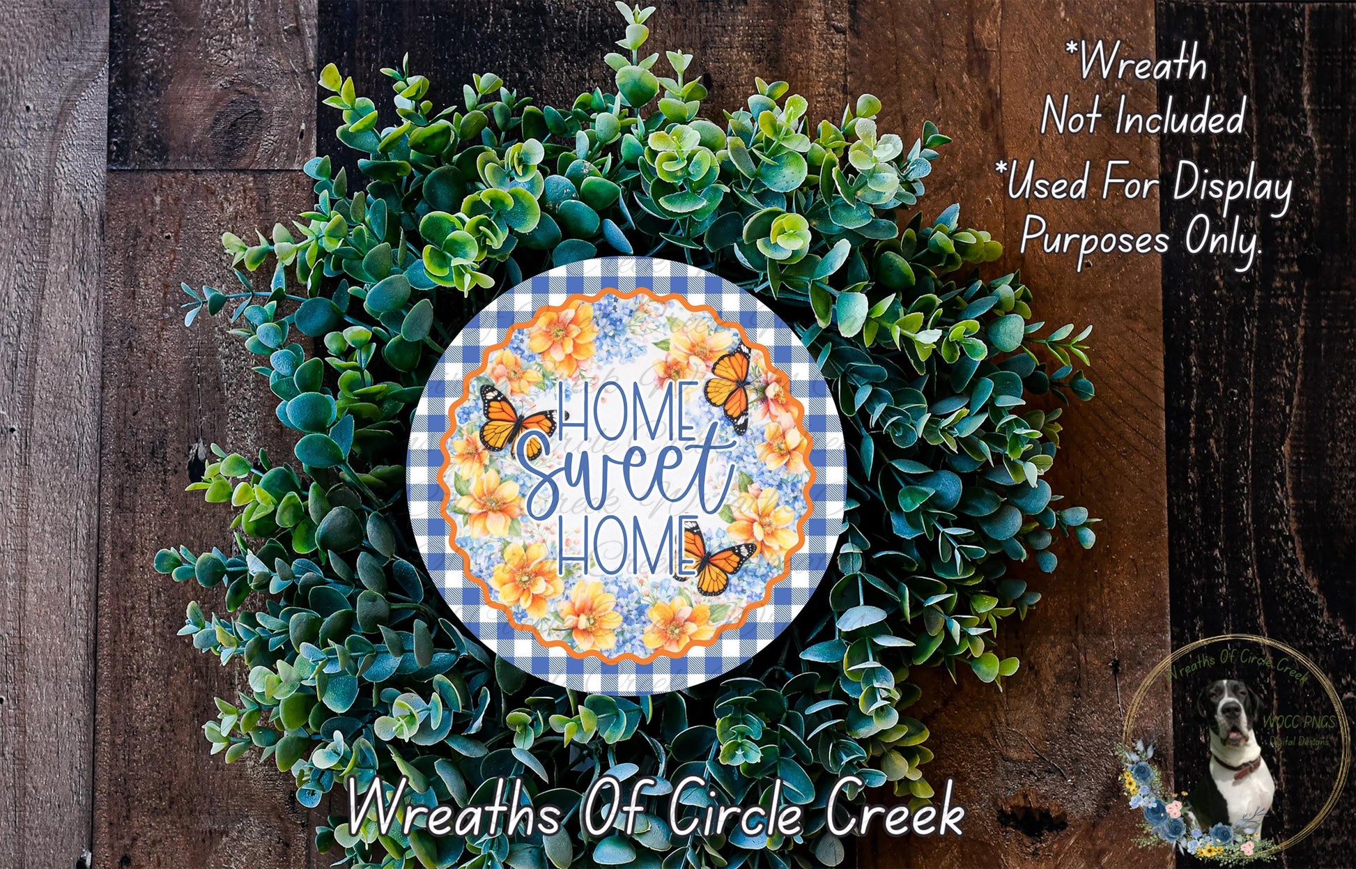 a wreath made of various green plants and flowers, with a decorative plate in the center that says "Home Sweet Home". The wreath is displayed on a wooden surface, and there is a small dog in the bottom right corner.