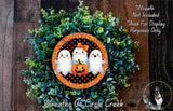 A Halloween-themed wreath made of green foliage and a black and orange border, with three small ghost-like figures holding a carved pumpkin in the center.