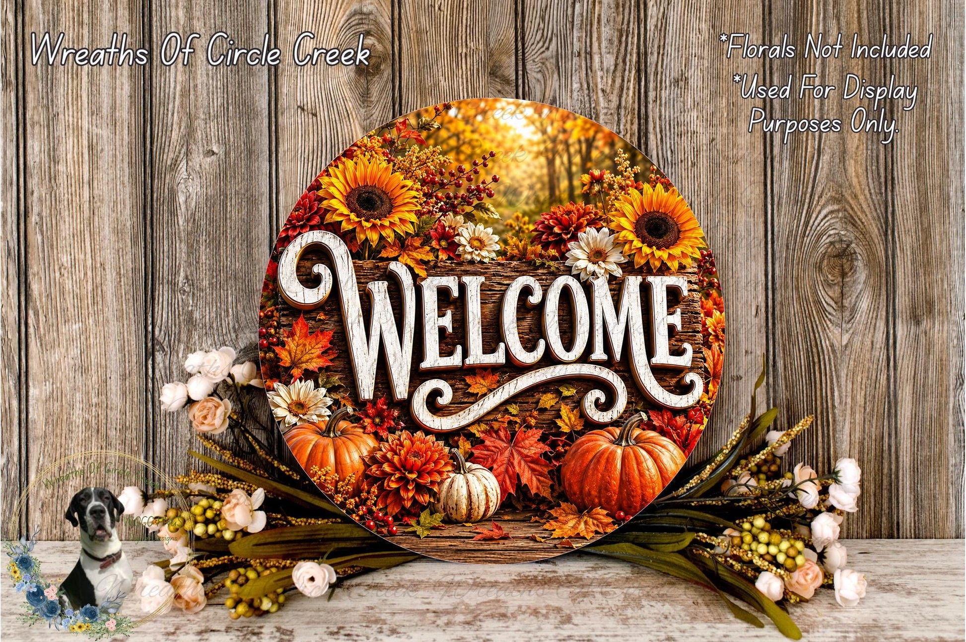 a circular welcome sign surrounded by autumn decorations, including sunflowers, pumpkins, and other seasonal elements. The sign is placed on a wooden surface, and there are flowers and foliage surrounding it.