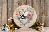 a round wooden plaque with a floral design and the word "Loved" written on it. The plaque is surrounded by a variety of flowers, including roses and other colorful blooms. The background is a wooden wall with a rustic, natural appearance.