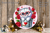A white cat wearing red glasses is sitting in a metal bucket, surrounded by red roses and a heart-shaped decoration.