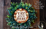 a wreath made of various green plants and flowers, with a circular sign in the center that says "YAY! you're here".