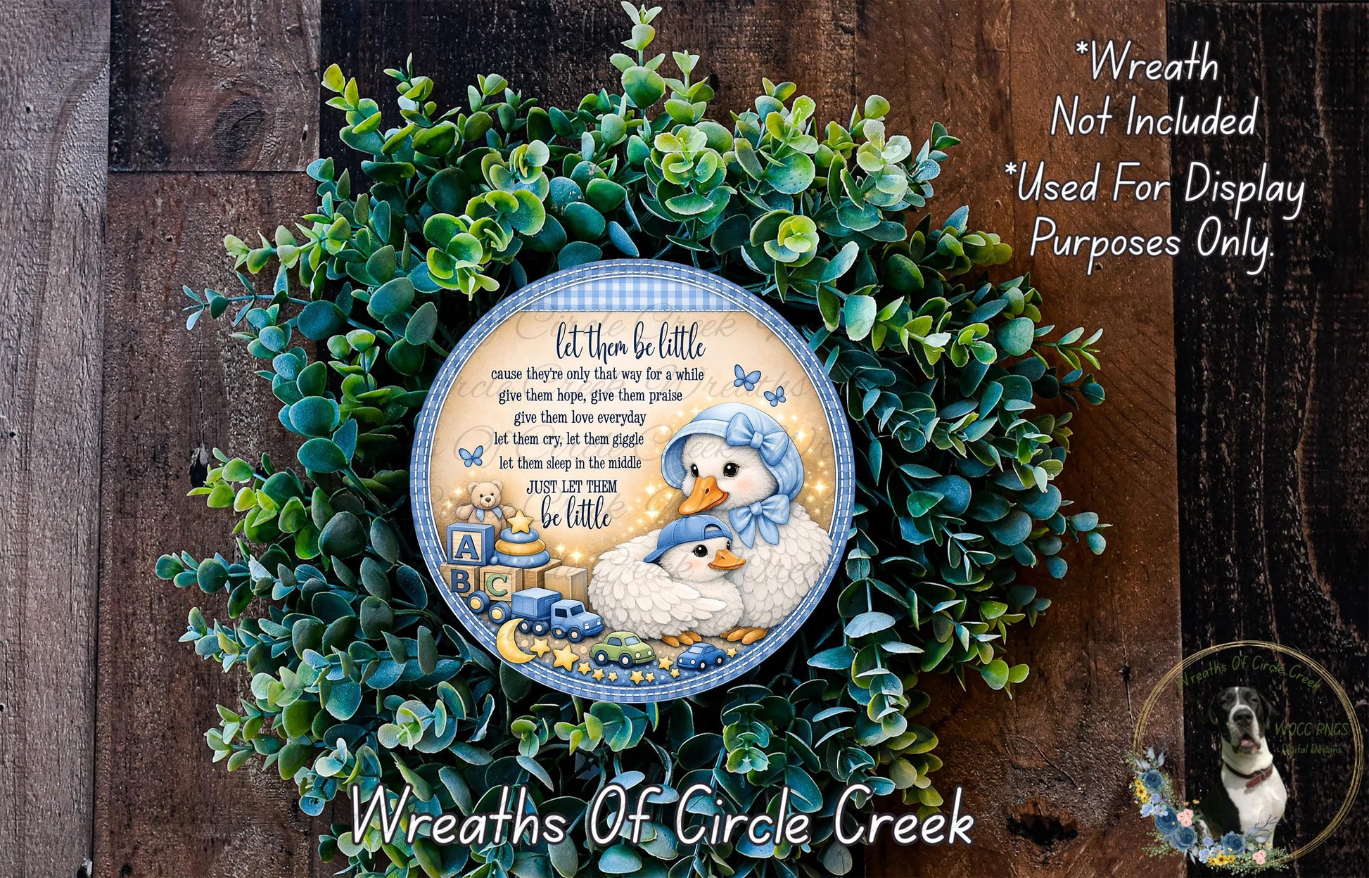 A wreath made of green leaves and featuring a blue and white plate with a cartoon image of two baby ducks. The wreath is displayed on a wooden surface, and there is a small dog in the bottom right corner.