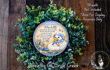 A wreath made of green leaves and featuring a blue and white plate with a cartoon image of two baby ducks. The wreath is displayed on a wooden surface, and there is a small dog in the bottom right corner.