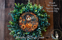 a Halloween-themed wreath with a pumpkin and a smiling jack-o-lantern. The wreath is surrounded by green foliage and has a circular plate with a pumpkin and the text "HELLO PHILL" on it. The wreath is displayed on a wooden surface, and there is a small dog in the bottom right corner.