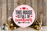 a round sign with the text "This house is full of sweethearts" and a heart symbol, surrounded by a floral arrangement on a wooden surface.
