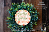 a wreath made of green leaves and a circular sign with the text "Sweeter than a Georgia peach" on it, placed on a wooden surface.