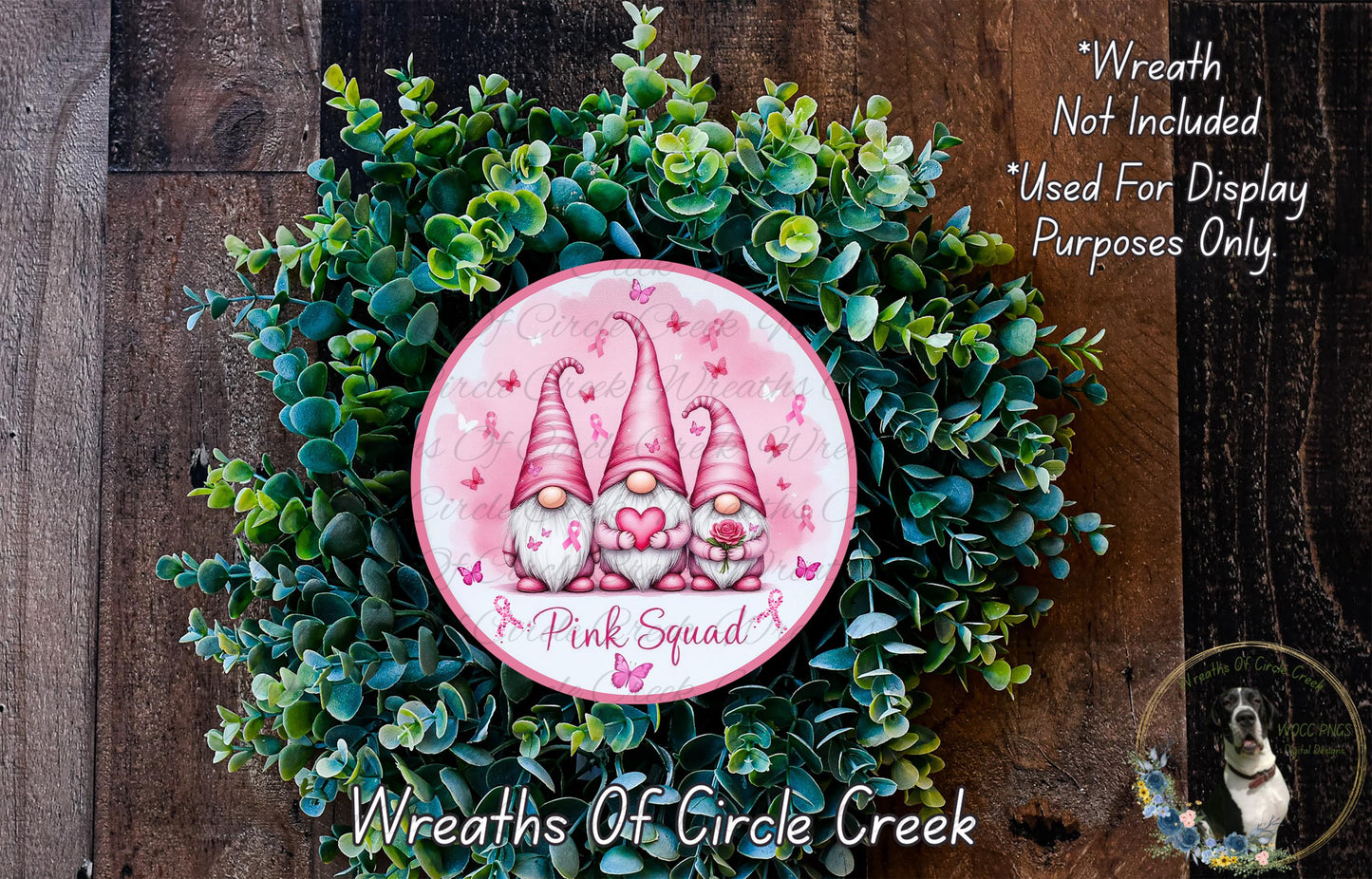 A wreath made of green leaves and flowers, featuring three pink gnomes holding a mushroom and a heart-shaped flower. The wreath is labeled "Pink Squad" and is displayed on a wooden surface.