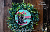 A wreath made of green leaves and flowers, with a pair of black and pink rain boots in the center, surrounded by the text "Hello Spring" and "Wreaths of Circle Creek".