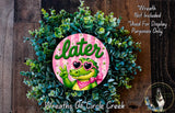 a wreath with a green alligator wearing sunglasses and a pink bow, surrounded by a lush green plant.