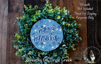 A wreath made of green leaves and flowers, with a blue circular sign that says "Happy Hanukkah" in the center.