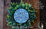 A wreath made of green leaves and flowers, with a blue circular sign that says "Happy Hanukkah" in the center.