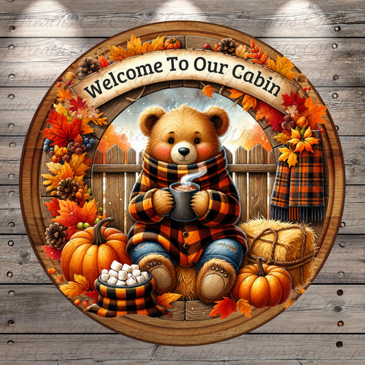 A wreath with a teddy bear wearing a scarf and holding a mug of hot chocolate, surrounded by autumn leaves, pumpkins, and hay bales, with the text "Welcome To Our Cabin" displayed above.
