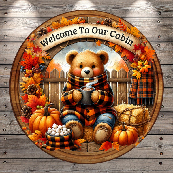 A wreath with a teddy bear wearing a scarf and holding a mug of hot chocolate, surrounded by autumn leaves, pumpkins, and hay bales, with the text &quot;Welcome To Our Cabin&quot; displayed above.
