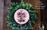 a wreath made of green leaves and a pink circular sign with the text "Jesus loves me, this I know" on it.