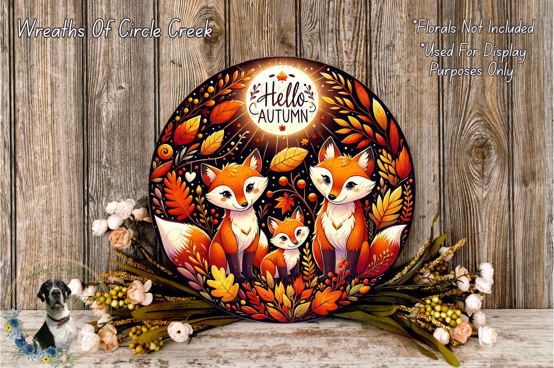 a decorative wreath with a central design of three foxes surrounded by autumn leaves and flowers. The wreath is placed on a wooden surface, and there are some flowers and leaves scattered around it.