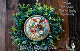 a wreath with a cow sitting in a bucket, surrounded by green foliage and flowers. The wreath is displayed on a wooden surface, and there is a small dog visible in the bottom right corner of the image.