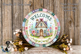 A decorative plate with a house and flowers, surrounded by a floral arrangement, against a wooden background.