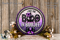 a decorative sign with the text "I put the BOO in BOUJEE" surrounded by a purple and black design, with a bow and flowers. The sign is placed on a wooden surface, and there are flowers and greenery in the foreground.