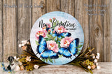 a round wooden plaque with a floral design, including blue butterflies and pink flowers. The plaque is placed on a wooden surface, surrounded by flowers and greenery.