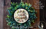 a wreath made of various green plants and a decorative plate with a pumpkin and other fall-themed items. The wreath is displayed on a wooden surface, and the plate is placed on the wreath.