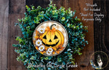 A wreath made of green foliage and a carved pumpkin with a carved face, placed on a wooden surface.