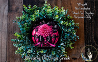 A wreath made of green leaves and a pink flower, with a black ribbon around it. The wreath is displayed on a wooden surface.