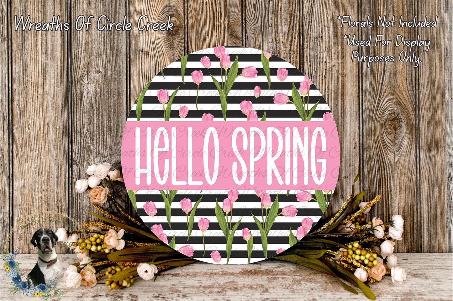 a round sign with the text "Hello Spring" in a pink color, surrounded by a floral arrangement of pink and white flowers. The sign is placed on a wooden surface, and there is a small dog in the background.