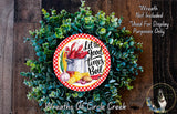 A wreath made of green foliage and featuring a red and white checkered border, with a circular design in the center displaying the text "Let the good times boil" surrounded by various food items such as a lobster, corn, and potatoes.