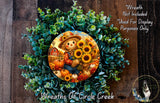 a wreath with a scarecrow and sunflowers, surrounded by greenery. The wreath is displayed on a wooden surface, and there is a small dog in the bottom right corner.