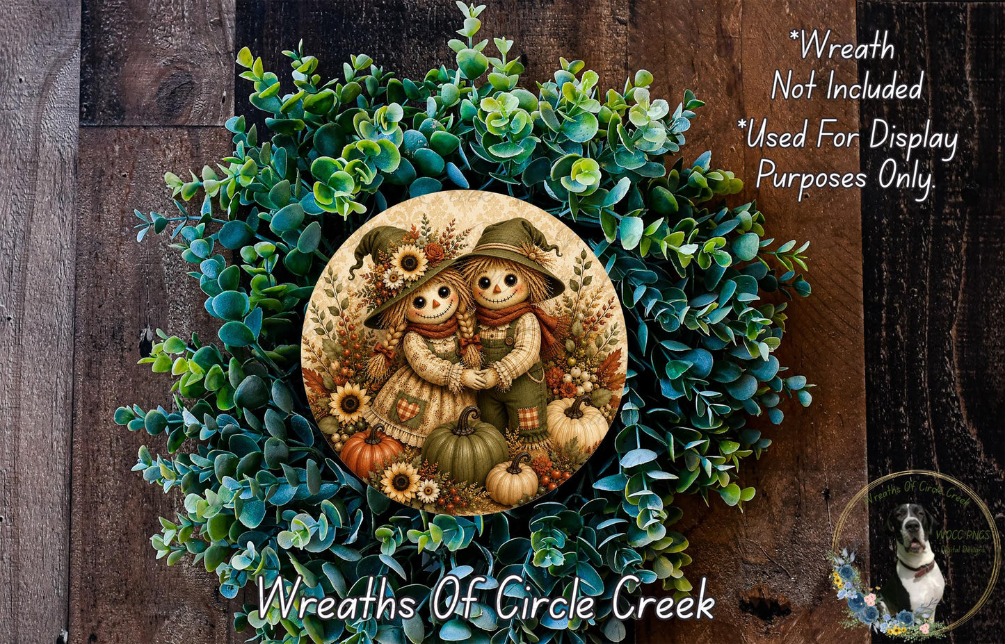 a wreath with a decorative design of two scarecrow figures, surrounded by various fall-themed elements such as pumpkins, leaves, and flowers. The wreath is placed on a wooden surface, and there is a small dog visible in the bottom right corner of the image.