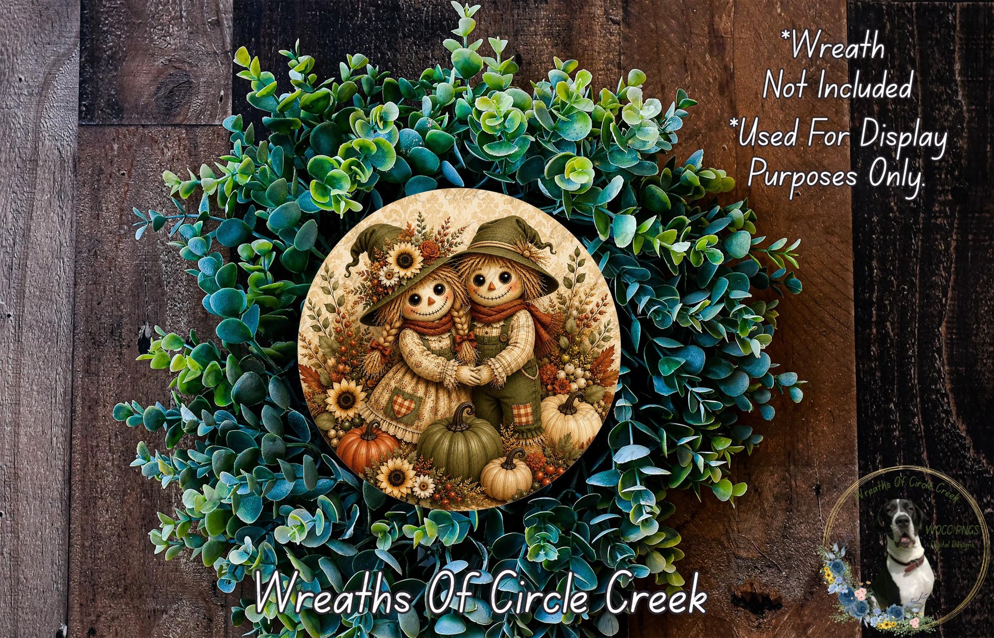 a wreath with a decorative design of two scarecrow figures, surrounded by various fall-themed elements such as pumpkins, leaves, and flowers. The wreath is placed on a wooden surface, and there is a small dog visible in the bottom right corner of the image.