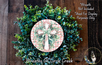 a wreath made of various green plants and flowers, including a pink and white checkered cross at the center. The wreath is displayed on a wooden surface, and there is a small dog visible in the bottom right corner of the image.