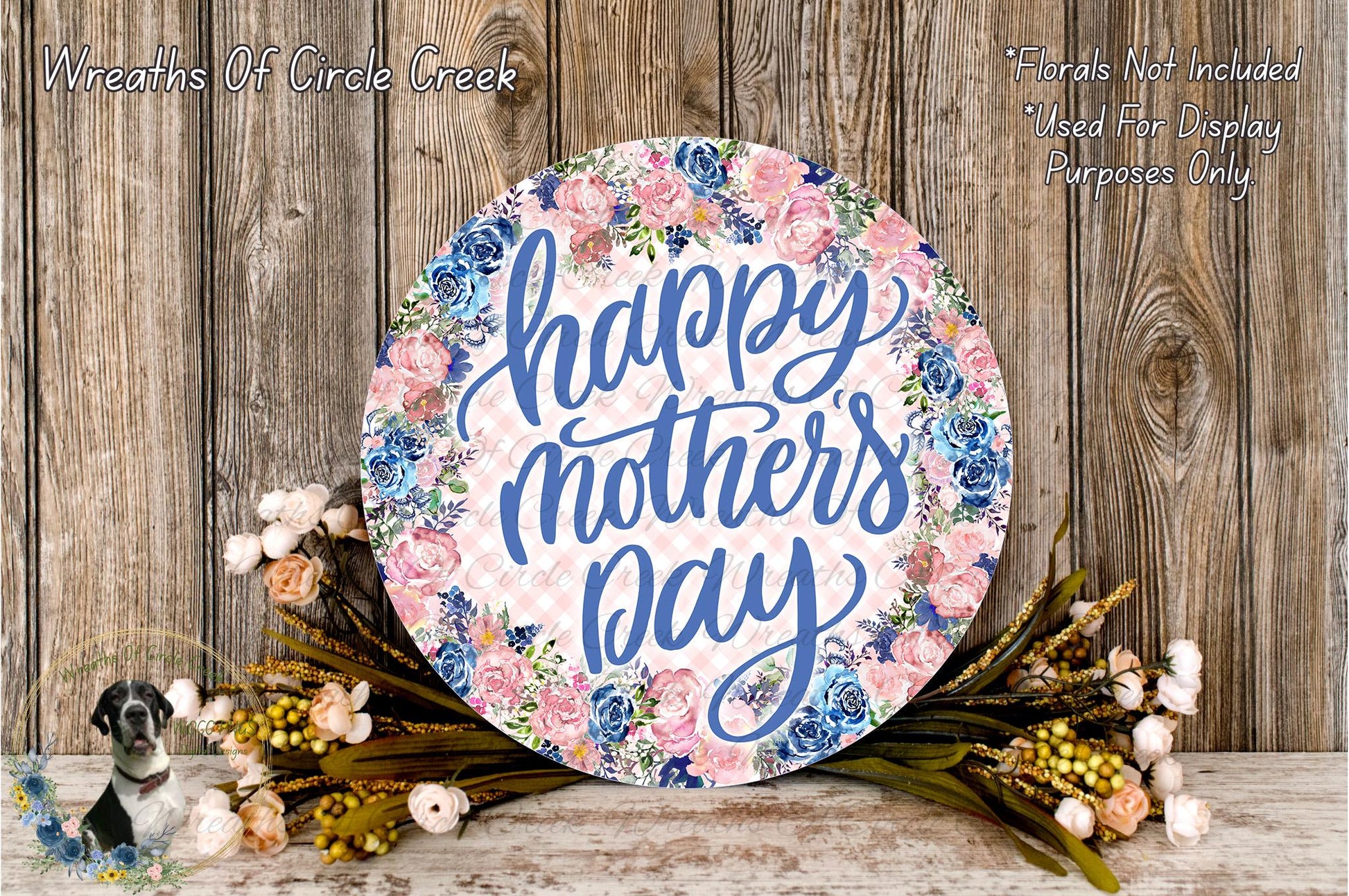 A round floral wreath with the text "Happy Mother's Day" written on it, surrounded by flowers and greenery, placed on a wooden surface.