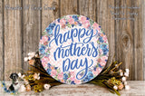 A round floral wreath with the text "Happy Mother's Day" written on it, surrounded by flowers and greenery, placed on a wooden surface.