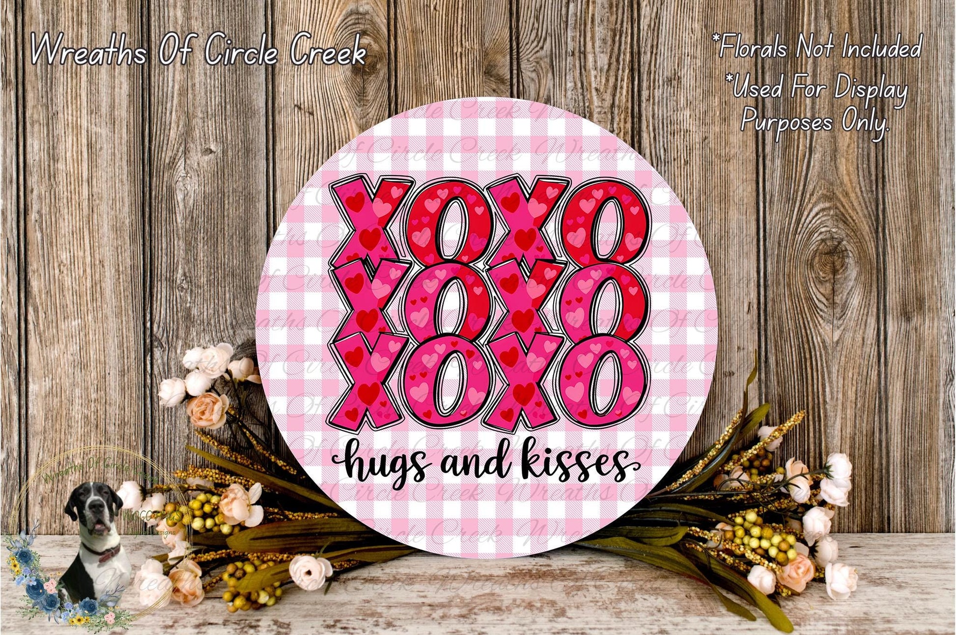 a round sign with the text "XOXO XOXO XOXO hugs and kisses" written on it, surrounded by a floral arrangement and a wooden background.