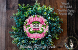 A wreath made of green leaves and flowers, with a pink and white circular sign that says "MERRY CHRISTMAS" in the center. The wreath is displayed on a wooden surface, and there is a text overlay that says "Wreath Not Included * Used For Display Purposes Only".