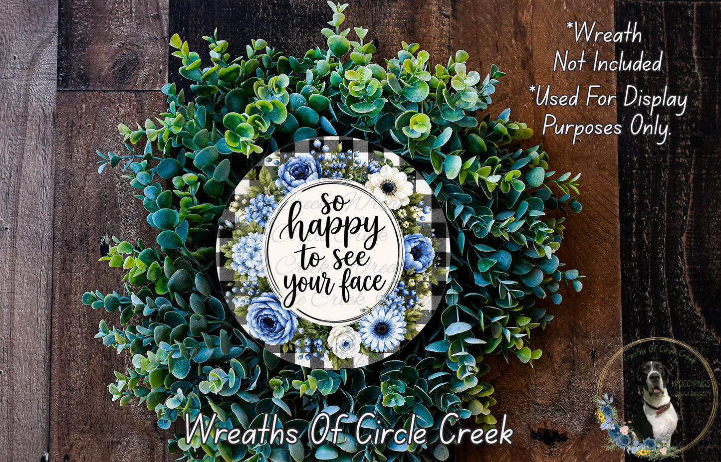 The image is a wreath made of various green and blue foliage, with a central circular design featuring a message that reads "So happy to see your face". The wreath is displayed on a wooden surface, and there is a small dog visible in the bottom right corner of the image.
