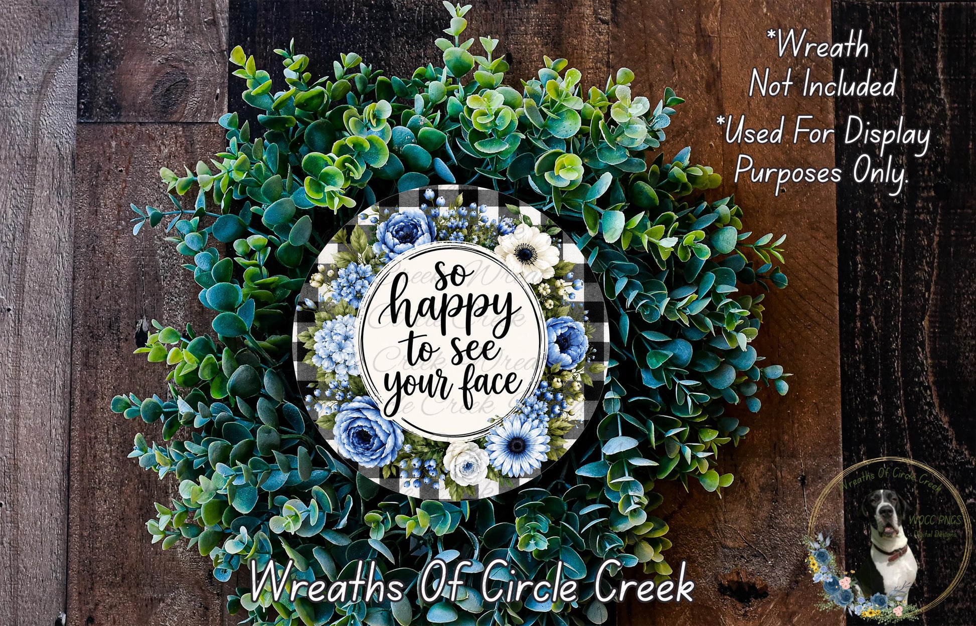 The image is a wreath made of various green and blue foliage, with a central circular design featuring a message that reads "So happy to see your face". The wreath is displayed on a wooden surface, and there is a small dog visible in the bottom right corner of the image.