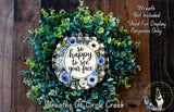 The image is a wreath made of various green and blue foliage, with a central circular design featuring a message that reads "So happy to see your face". The wreath is displayed on a wooden surface, and there is a small dog visible in the bottom right corner of the image.