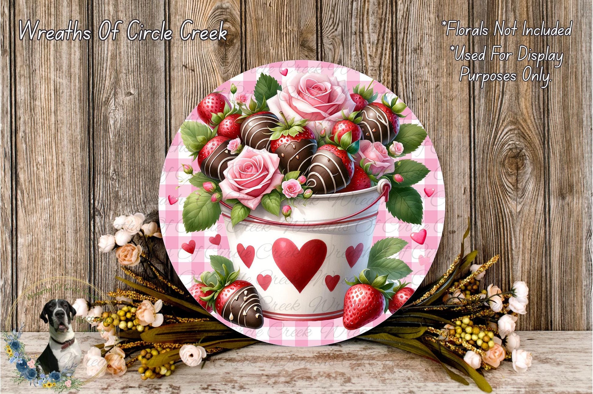 A decorative arrangement featuring a white bucket with a red heart and heart-shaped chocolate-covered strawberries, surrounded by a floral arrangement and a wooden background.